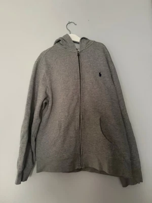 Grå hoodie med dragkedja från Polo Ralph Lauren - Klassisk grå hoodie från Polo Ralph Lauren med dragkedja framtill, två fickor och broderad logga på bröstet. Mjuk bomullsblandning och huva gör den perfekt för chill dagar. Enkel och stilren design som funkar till det mesta.