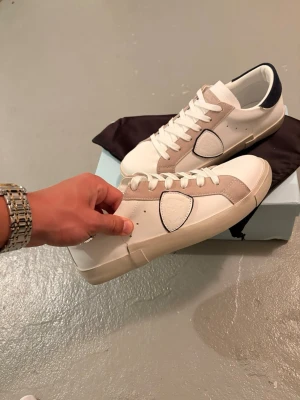 Philippe Model vita sneakers med mockadetaljer - Snygga vita sneakers från Philippe Model med beige mockadetaljer och mörkblå häl. Skorna har klassisk låg siluett, vita skosnören och en ikonisk sköldlogga på sidan. Tillverkade i läder och mocka för en lyxig känsla.