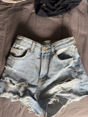 Bershka Ljusa denimshorts  - Säljer ett par ljusblå denimshorts från Bershka med hög midja och råa, fransiga kanter. Shortsen har flera slitningar framtill och bakfickor med distressed detaljer. Perfekta för sommaren och ger en avslappnad vibe.