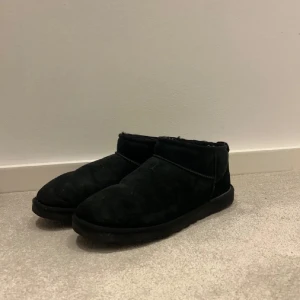 Svarta UGG boots i mocka - Säljer ett par svarta UGG boots med låg skaft och rund tå. Skorna är tillverkade i mjuk mocka med fluffigt foder på insidan för extra värme. Klassisk UGG-logga baktill och platt sula som ger skön komfort. Perfekt för kalla dagar.