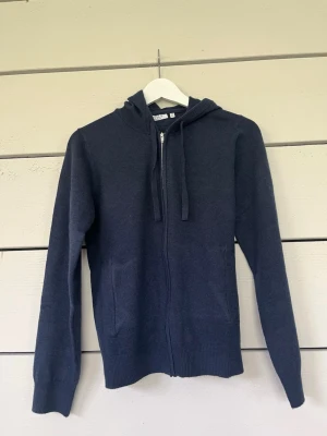 Mörkblå stickad hoodie med dragkedja - Mörkblå stickad hoodie från RYVLS med dragkedja framtill, två fickor och justerbar huva med snören. Tröjan har ribbade muddar vid ärmslut och nederkant. Perfekt för en avslappnad och stilren look.