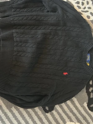 Svart kabelstickad tröja Polo Ralph Lauren - Kabelstickad svart tröja från Polo Ralph Lauren med klassisk rund halsringning och röd broderad logga på bröstet. Tröjan har ribbstickade muddar och är perfekt för en stilren look.
