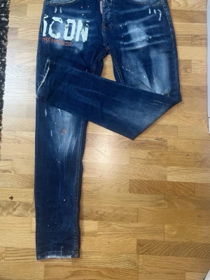 Mörkblå Dsquared2 ICON jeans - Säljer ett par mörkblå Dsquared2 ICON jeans med vit logga och tryck på vänster lår. Jeansen har slitningar, färgstänk och en tvättad look för en trendig och avslappnad stil. Smal passform och klassisk femficksmodell. Materialet är denim i bomull.