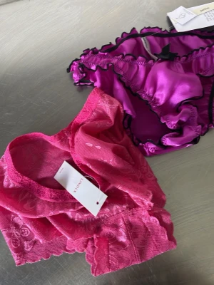 Två par färgglada trosor från Lindex - Säljer två par snygga trosor från Lindex. Ett par är rosa i spets med blommönster och bredare modell, det andra paret är lila i glansigt material med svarta volangkanter och liten rosett framtill. Perfekta för dig som gillar färg och fina detaljer.