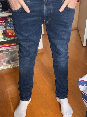 Mörkblå slim nudie jeans - Snygga mörkblå slim jeans med klassisk femficksdesign och guldfärgade sömmar. Jeansen har en smal passform och är tillverkade i stretchigt denimtyg för extra komfort. Perfekta för en trendig och avslappnad stil. Nypris 1600kr