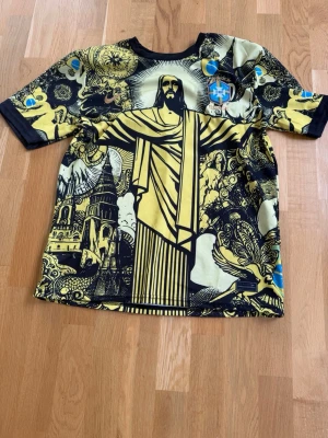 Gul Brasilien t-shirt med grafiskt tryck - Unik t-shirt med stort grafiskt tryck av Kristusstatyn och brasilianska motiv i svart och gult. Rund halsringning och korta ärmar. Detaljer i blått och vitt samt Brasil-logga på bröstet. Perfekt för dig som vill sticka ut med en färgstark och konstnärlig look.