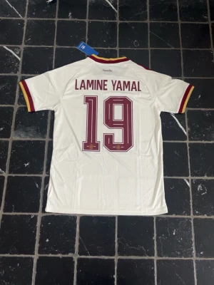 Spanien Lamine Yamal matchtröja Adidas - Vit Spanien fotbollströja från Adidas med Lamine Yamal och nummer 19 på ryggen. Tröjan har röda och gula detaljer vid ärmslut och krage, samt röda Adidas-ränder på axlarna. Broderad landslagssköld och Adidas-logga framtill. Perfekt för fotbollsfans!