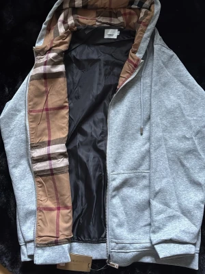 Grå hoodie med dragkedja från Burberry - Snygg grå hoodie från Burberry med dragkedja och klassiskt rutigt foder i huvan. Hoodien har dragsko i huvan, ribbade muddar och två fickor framtill. Perfekt för en chill och stilren look. Materialet är mjukt och bekvämt, med exklusiva detaljer som sticker ut.