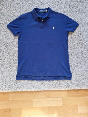 Polo Ralph Lauren marinblå pikétröja  - Marinblå Polo Ralph Lauren pikétröja med gul logga i storlek M, passar L också. Custom slim fit. 100% bomull. Har nästan inte använt den, så den är som ny. Inga defekter eller slitningar finns. Passar perfekt för dig som vill vara stilig under sommaren! Skick: 9.5/10