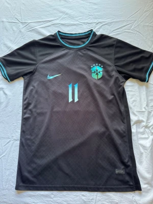 Svart Brasilien Nike fotbollströja - Snygg svart fotbollströja från Nike med Brasilien-emblem och nummer 11 i turkos på bröstet. 'Romário' och nummer 11 på ryggen. Perfekt för dig som älskar fotboll och vill sticka ut på planen. Kan gå ner vid snabb affär.