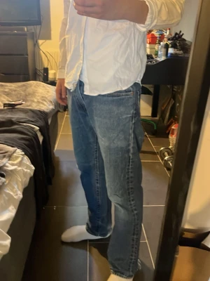 Blå raka jeans i klassisk modell - Säljer ett par blå jeans med rak passform och klassisk femficksdesign. Jeansen har normalhög midja och är tillverkade i denim med synliga sömmar. Perfekta för en avslappnad och stilren look. Har defekter längst ner på byxorna.