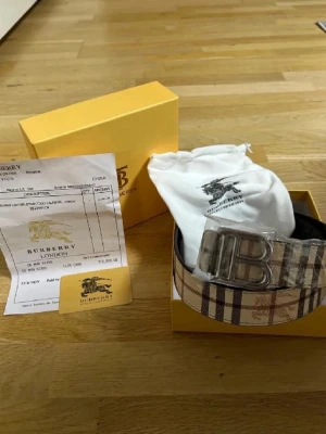 Burberry rutigt bälte med silverdetalj - Snyggt Burberry-bälte med klassiskt rutigt mönster i beige, svart och rött. Bältet har ett stort silvrigt spänne med Burberrys logotyp och detaljer i skinn. Perfekt accessoar för att lyfta din outfit.