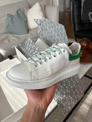 Alexander McQueen skor - Snygga vita Alexander McQueen skor med tjock sula och grön krokodil detalj vid hälen. Skorna har vita och gröna snören samt perforerade hål på sidorna för ventilation. Tillverkade i slätt lädermaterial och har en klassisk låg profil som passar till många stilar.