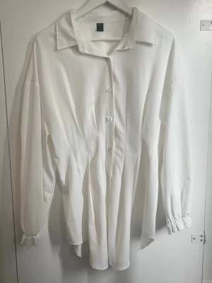 Vit oversized skjorta - Klassisk vit skjorta från med figursydd passform. Skjortan har långa ärmar med lätt rynkade muddar, knappar framtill och en bred krage. Perfekt för en stilren och enkel look.