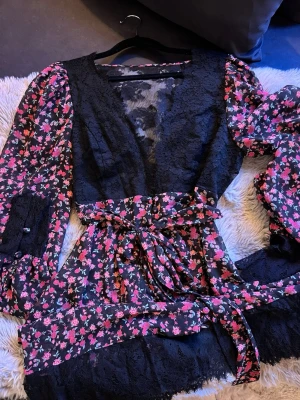 Blommig omlottklänning For Love & Lemons - Supersöt kort omlottklänning från For Love & Lemons x Victoria’s secret med svart spets upptill och blommigt mönster i rosa och rött. Klänningen har långa ärmar, knyt i midjan och volangdetaljer. Perfekt för dig som gillar romantisk och trendig stil. Helt ny, aldrig använd
