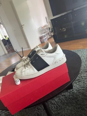 Valentino Garavani vita sneakers - Snygga vita sneakers från Valentino Garavani med en bred svart rem över sidan och coola nitar på sulan. Skorna har klassisk låg profil, vita snören och detaljerad logga på plösen. Perfekta för dig som vill sticka ut med stilrena och exklusiva sneakers. Allt og med följer 