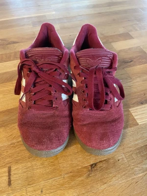 Vinröda Adidas Busenitz sneakers - Snygga vinröda Adidas Busenitz sneakers med klassiska vita ränder på sidorna och gummisula. Ovandelen är i mocka och skon har lågt skaft. Perfekta för dig som gillar streetstyle och vill ha en ikonisk look. Storlek 42/5. Kan diskutera om priset