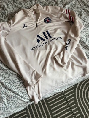 PSG träningströja beige långärmad - Snygg beige långärmad träningströja från PSG och Jordan med dragkedja vid halsen. Tröjan har mörkblå logga och text, röda och vita ränder på ena ärmen samt tryck på ärmen och ryggen. Perfekt för fotbollsträning eller chill.