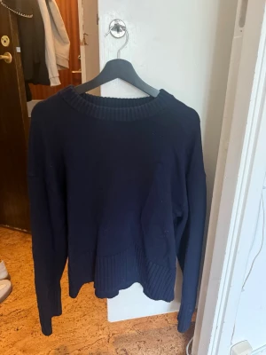 Mörkblå stickad tröja från H&M - Mysig mörkblå stickad tröja från H&M med bred ribbad kant vid halsringning, ärmslut och nederkant. Tröjan har en avslappnad passform och är perfekt för lager-på-lager-stil. Materialet är mjukt och bekvämt, vilket gör den enkel att bära hela dagen.