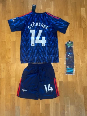 Arsenal bortaställ blå Gyökeres #14 - Säljer ett Arsenal bortaställ från Adidas med Gyökeres #14. Tröjan är blå med blixtmönster, röda ränder på axlarna och klubbmärke på bröstet. Setet inkluderar matchtröja, shorts och strumpor. Materialet är lätt och andas, perfekt för fotbollsträning.