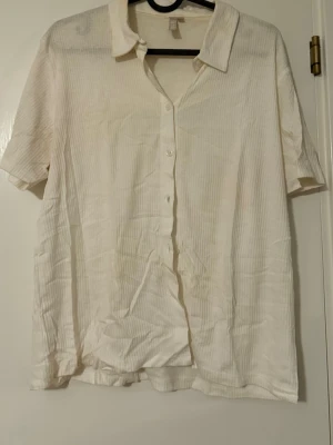 Vit kortärmad blus från Pieces - Vit blus från Pieces med korta ärmar och klassisk krage. Blusen har knappar framtill och är tillverkad i ett lätt, tunt tyg med diskret randig struktur. Perfekt för en avslappnad och stilren look.