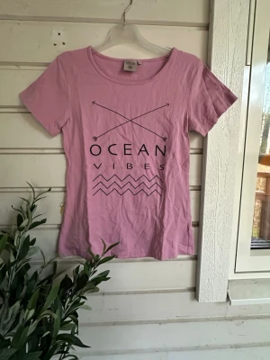 Rosa t-shirt med tryck Ocean Vibes storlek: M - Snygg rosa t-shirt från Vila med svart tryck som säger 'Ocean Vibes' och har två korsade pilar samt vågmönster. T-shirten har rund halsringning och korta ärmar. Perfekt för en avslappnad stil.