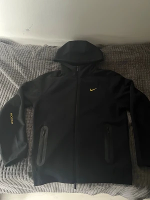Svart Nike NOCTA hoodie med dragkedja - Svart hoodie från Nike NOCTA med hel dragkedja, huva och två fickor med dragkedja. Liten gul Nike-logga på bröstet och NOCTA-tryck på ärmen. Snygg och stilren design, perfekt för streetwear. Materialet känns mjukt och sportigt.