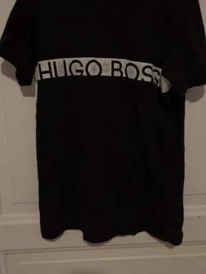 Svart Hugo Boss t-shirt med logga - Svart t-shirt från Hugo Boss med vit logga tvärs över bröstet. Klassisk passform och korta ärmar, perfekt för en stilren och enkel look. Tillverkad i mjuk bomull som känns skön mot huden. Strlk M (passar bättre på M)