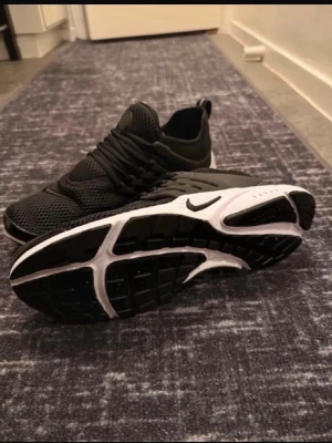 Svarta Nike Air Presto sneakers - Snygga svarta Nike Air Presto sneakers med vit och svart sula. Ovandelen är i mesh för bra andningsförmåga och skon har en sportig, strömlinjeformad design med klassisk Nike-logga på sidan. Perfekta för en streetwear-look.