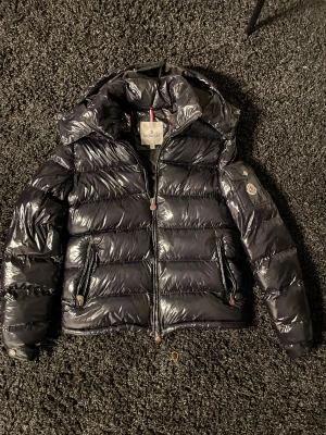 Svart Moncler pufferjacka M - Säljer en svart pufferjacka från Moncler i storlek M. Jackan har glansig finish, huva, två sidofickor med dragkedja och en ficka med Moncler-logga på ärmen. Jackan har tyvär fått en liten reva på högra armen men det är lagat med svart tejp. Jackan är bara använd ett fåtal gånger så den är så gott som ny.
