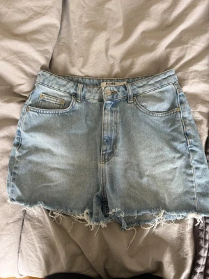 Ljusblå jeansshorts från NA-KD - Säljer ett par ljusblå jeansshorts från NA-KD med råa, fransiga benslut och klassisk femficksdesign. Shortsen har hög midja och är tillverkade i bomull för en skön och avslappnad look. Perfekta för varma dagar och enkel att styla med allt.