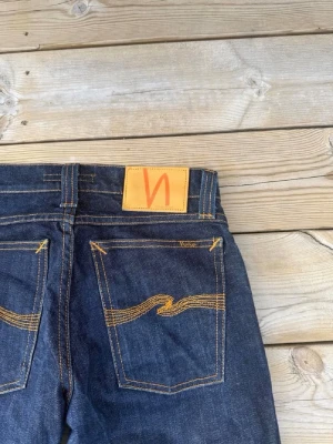 Mörkblå jeans från Nudie Jeans - Säljer ett par mörkblå jeans från Nudie Jeans med klassisk rak passform. Jeansen har kontrastsömmar i gult och Nudie-loggan på bakfickan samt läderpatch i midjan. Tillverkade i kraftig denim, perfekta för en avslappnad och stilren look.