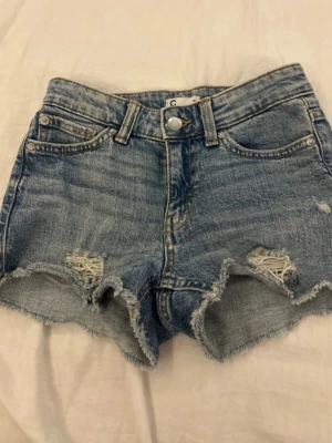 Blåa högmidjade jeansshorts - Snygga blå jeansshorts med hög midja och råa, fransiga kanter. Shortsen har slitna detaljer framtill och klassisk femficksdesign. Perfekta för sommaren och ger en avslappnad vibe.