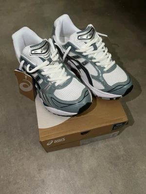 ASICS vita och grå sneakers - Snygga sneakers från ASICS i vitt och grått med svarta detaljer. Ovandelen är i mesh och syntetmaterial för bra andningsförmåga och komfort. Klassisk snörning och sportig design som passar perfekt till streetwear och avslappnade outfits.