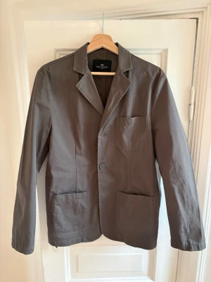  John Henric overshirt  - Stilren grå kavaj från John Henric med tre utanpåliggande fickor och klassisk krage. Tillverkad i ett lätt bomullstyg som ger en avslappnad men snygg look. Perfekt för dig som gillar enkel och modern stil.