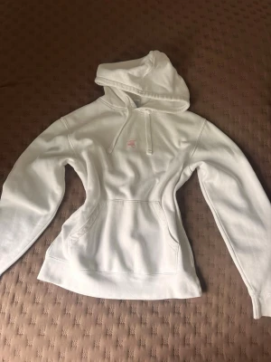 Vit hoodie från Russell - Klassisk vit hoodie från Russell med huva och dragsko. Hoodien har en stor magficka och är tillverkad i mjukt och bekvämt material, perfekt för en avslappnad stil. Enkel design med liten logga på bröstet och ribbade muddar vid ärmslut och nederkant.