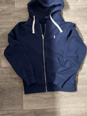  Ralph Lauren zip hoodie  - Snygg marinblå hoodie med dragkedja från Polo Ralph Lauren. Klassisk design med vit broderad logga på bröstet, vita snören i huvan och två stora fickor framtill. Tillverkad i mjukt bomullsmaterial och har ribbade muddar vid ärmslut och nederkant.