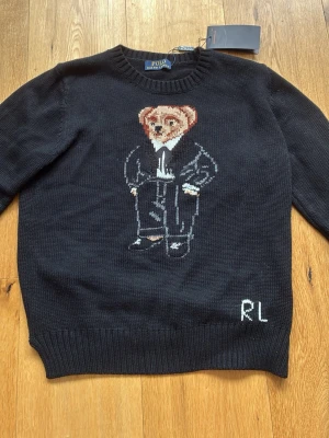 Svart stickad tröja Polo Ralph Lauren - Svart stickad tröja från Polo Ralph Lauren med ikonisk björn i kostym på bröstet och RL-broderi nedtill. Tröjan har rund halsringning och ribbade muddar. Perfekt för dig som gillar klassiska och lekfulla detaljer. Valde att sälja den på grund av att den var för liten när jag köpte den. Aldrig använd. Hör av er om ni har funderingar, kan diskutera pris