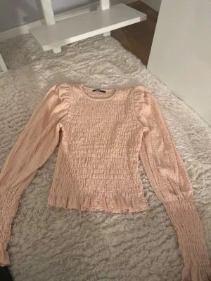 Ljusrosa smockad blus med volang - Supersöt ljusrosa blus med smockat tyg och volangkant nertill. Blusen har långa ärmar med lätt puff vid axlarna och rund halsringning. Perfekt för en trendig och feminin look. Jätte bra skick. De är en storlek L men tycker den passar en storlek M också.