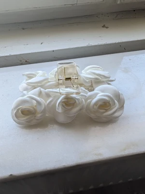 Vit hårklämma med tygrosor - Superfin hårklämma i transparent plast dekorerad med vita tygrosor. Perfekt accessoar för att ge en romantisk och söt touch till din frisyr. Klämman har en stabil fjäder och rosorna är mjuka och fluffiga.