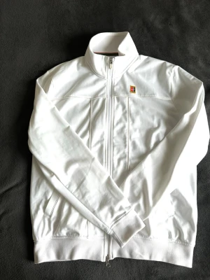 Vit träningsjacka från Nike - Snygg vit träningsjacka från Nike med hel dragkedja, ståkrage och två fickor framtill. Jackan har en liten färgglad logotyp på bröstet och är tillverkad i ett mjukt, sportigt material som passar perfekt till träning eller uppvärmning.
