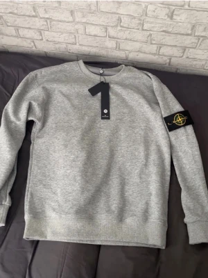 Grå sweatshirt från Stone Island - Snygg grå sweatshirt från Stone Island med klassisk rund hals och ribbade muddar. Tröjan har den ikoniska svarta logotypen med kompass på vänster ärm. Tillverkad i mjuk bomullsblandning för en bekväm känsla.