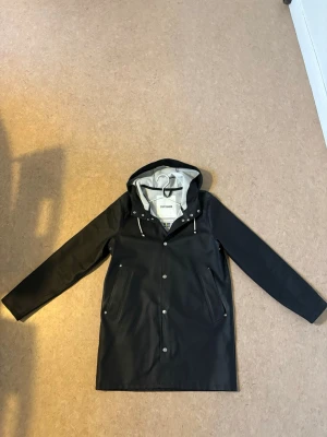 Svart regnjacka från Stutterheim - Snygg svart regnjacka från Stutterheim med huva och vita dragsnören. Storlek XS, funkar även med S Väldigt fint och hållbart märke. Jackan har tryckknappar framtill, två fickor och är fodrad med ljusgrått tyg. Nypris 3000kr Perfekt för regniga dagar och stilren look. Inga slitage. Andvänt 5 gånger