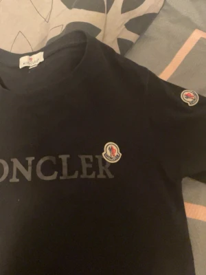 Svart Moncler t-shirt med logga - Snygg svart t-shirt från Moncler med stor logga på bröstet och en mindre patch på ärmen. Klassisk rund hals och korta ärmar. Tillverkad i mjuk bomull som känns skön mot huden. Perfekt för dig som gillar stilrena och exklusiva plagg. Pris kan diskuteras