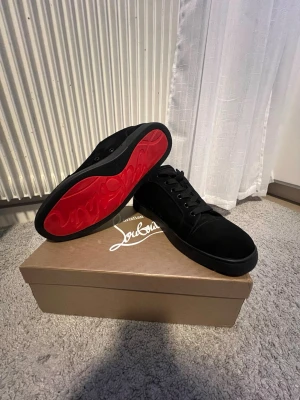 Svarta Christian Louboutin sneakers - Snygga svarta sneakers från Christian Louboutin med klassisk röd sula och diskret logga. Skorna har en låg profil, rund tå och är tillverkade i mocka med svarta skosnören. Perfekta för dig som vill ha en lyxig och stilren look.