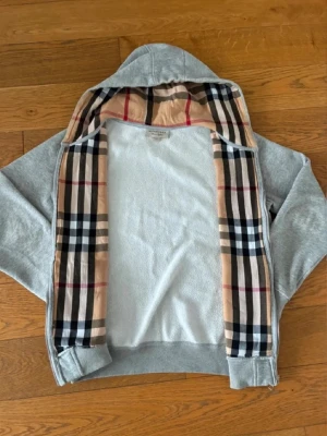 Grå zip hoodie från Burberry - Säljer en grå zip hoodie från Burberry med klassiskt rutigt foder i huvan och längs dragkedjan. Hoodien har två fickor framtill, ribbade muddar och är tillverkad i mjukt bomullsmaterial. Perfekt för en chill och stilren look.