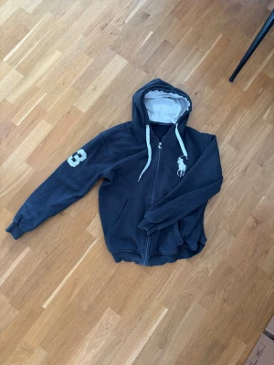 Svart hoodie från Polo Ralph Lauren - Snygg svart hoodie med dragkedja från Polo Ralph Lauren. Stor vit logga på bröstet och siffran 3 på ärmen. Vit insida i huvan och vita dragsnören ger en schysst kontrast. Perfekt för en avslappnad och sportig stil.