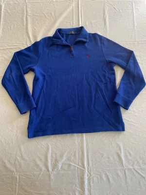 Blå halvzip från Polo Ralph Lauren - Snygg blå långärmad tröja från Polo Ralph Lauren med halv dragkedja och liten röd logga på bröstet. Tröjan har hög krage och är tillverkad i mjukt material, perfekt för lager-på-lager-stil.   Tveka inte, hör av er vid intresse, frågor eller prisförslag!