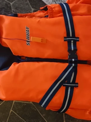 Orange flytväst från Seaway - Säljer en orange flytväst från Seaway med svarta och vita justerbara remmar och spänne framtill. Västen har visselpipa och är designad för att ge extra säkerhet på vattnet. Perfekt för båtturer eller andra vattenaktiviteter.