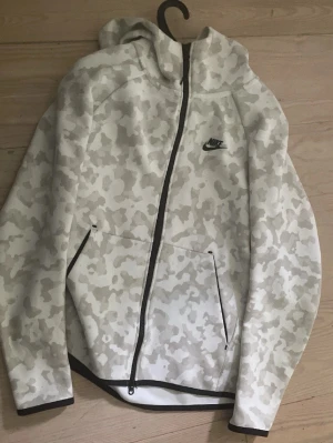 Nike Tech med camomönster - Snygg hoodie från Nike i vitt och ljusgrått camomönster. Dragkedja framtill, två fickor och svart Nike-logga på bröstet. Huva och svarta detaljer vid dragkedja och muddar. Perfekt för en sportig och avslappnad stil.